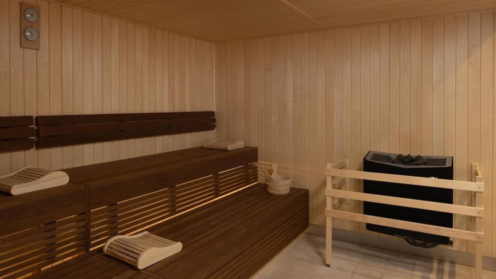 Wood-paneled hotel sauna with tiered wooden benches, folded towels, sauna heater and temperature gauges.