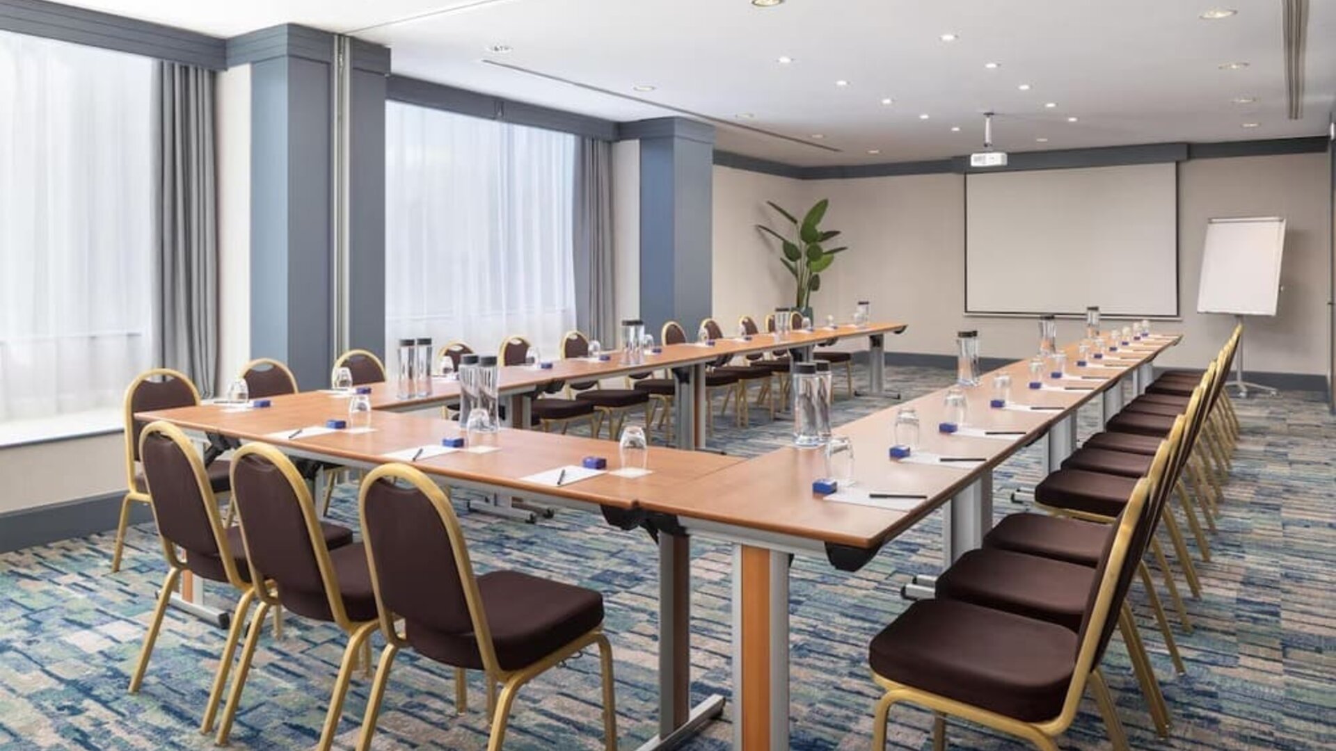 Hotel meeting room with U-shaped conference tables, chairs, presentation screen, flip chart and bottled water.