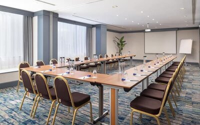 Hotel meeting room with U-shaped conference tables, chairs, presentation screen, flip chart and bottled water.