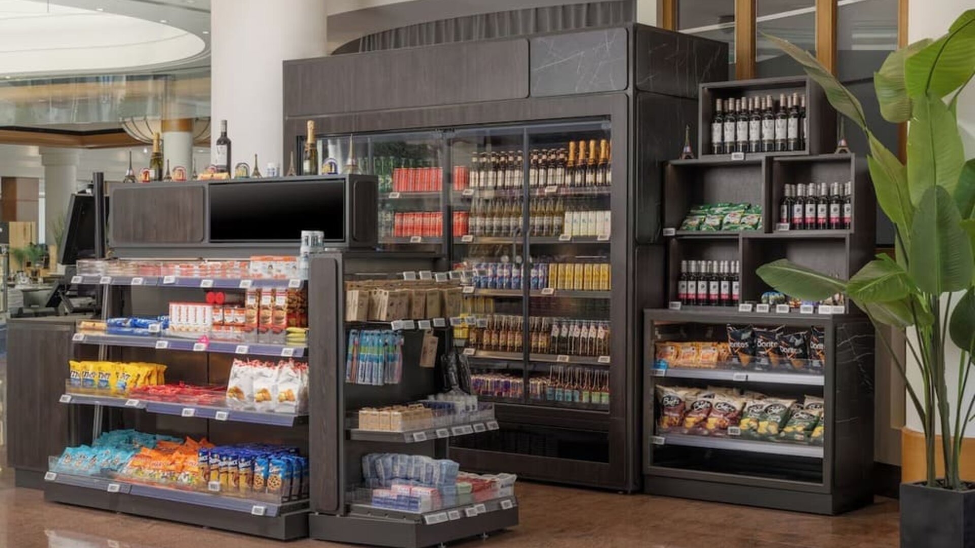 Hotel convenience store with refrigerated drinks, snacks, wine, toiletries and travel essentials displayed on shelves.