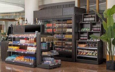 Hotel convenience store with refrigerated drinks, snacks, wine, toiletries and travel essentials displayed on shelves.