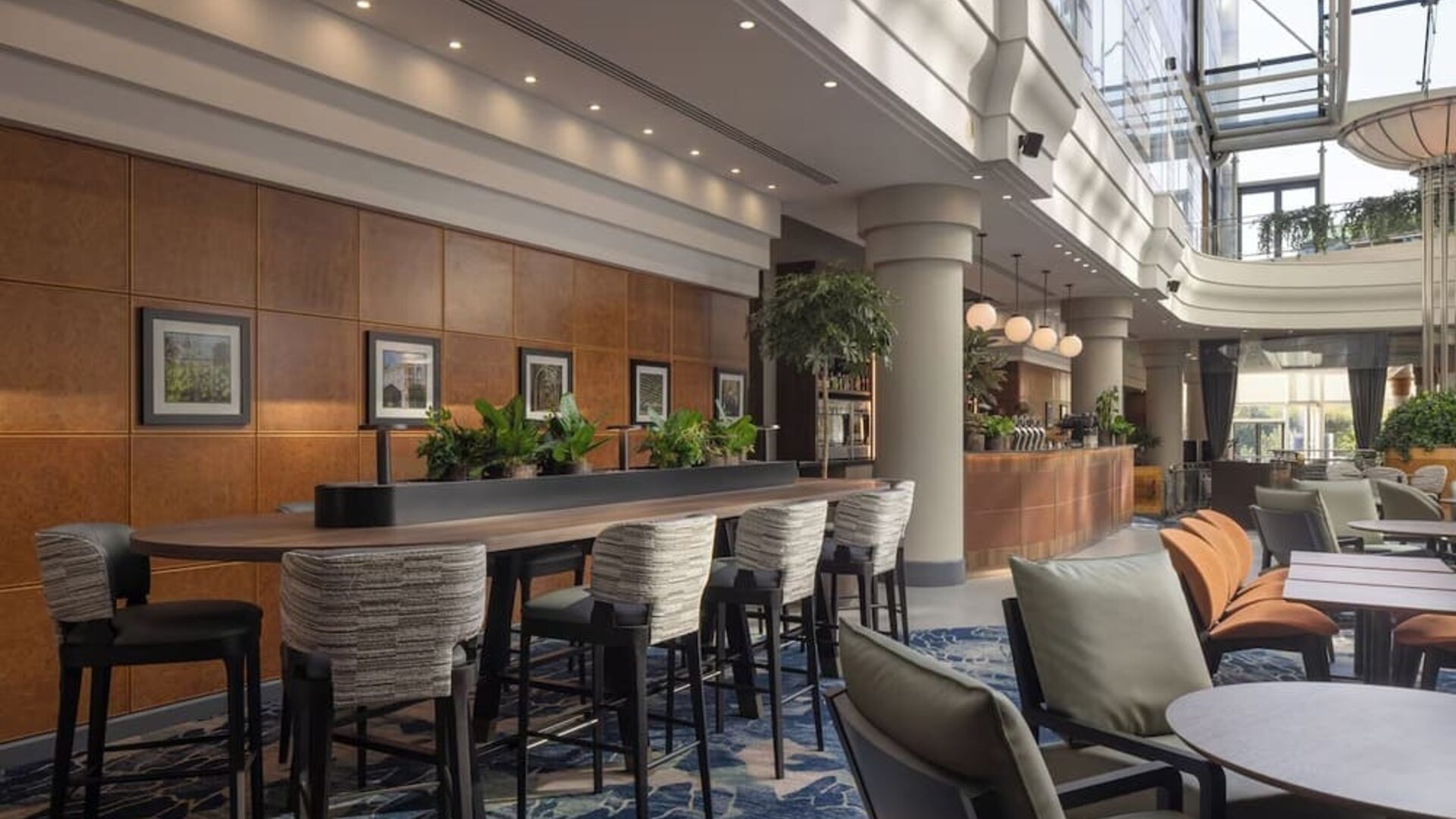 Bright hotel coworking and lounge area with communal table, high chairs, comfortable seating and a glass atrium.