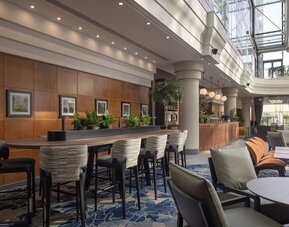 Bright hotel coworking and lounge area with communal table, high chairs, comfortable seating and a glass atrium.