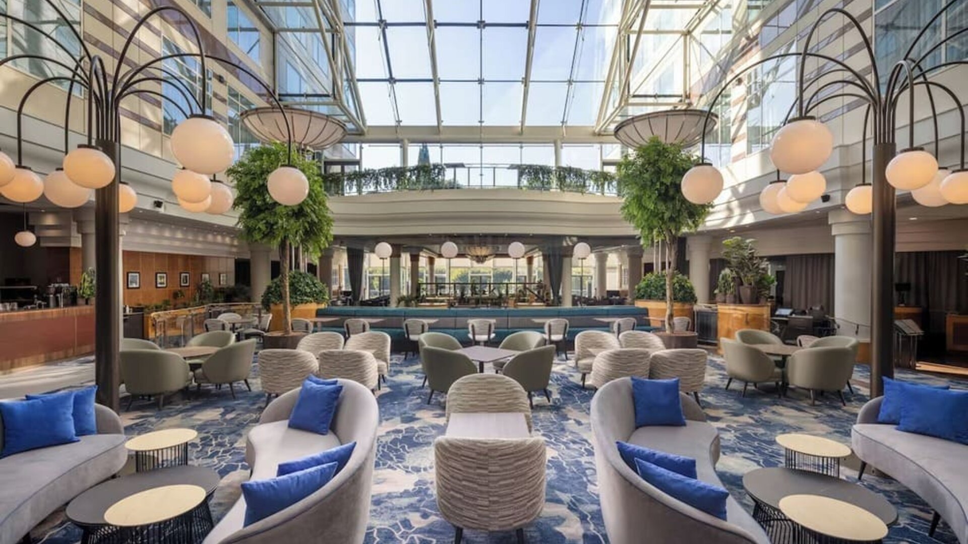 Expansive hotel lobby lounge beneath a glass atrium with sofas, armchairs, tables, blue carpet and globe lighting.
