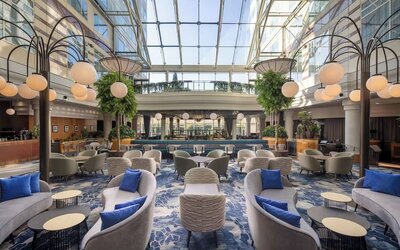 Expansive hotel lobby lounge beneath a glass atrium with sofas, armchairs, tables, blue carpet and globe lighting.