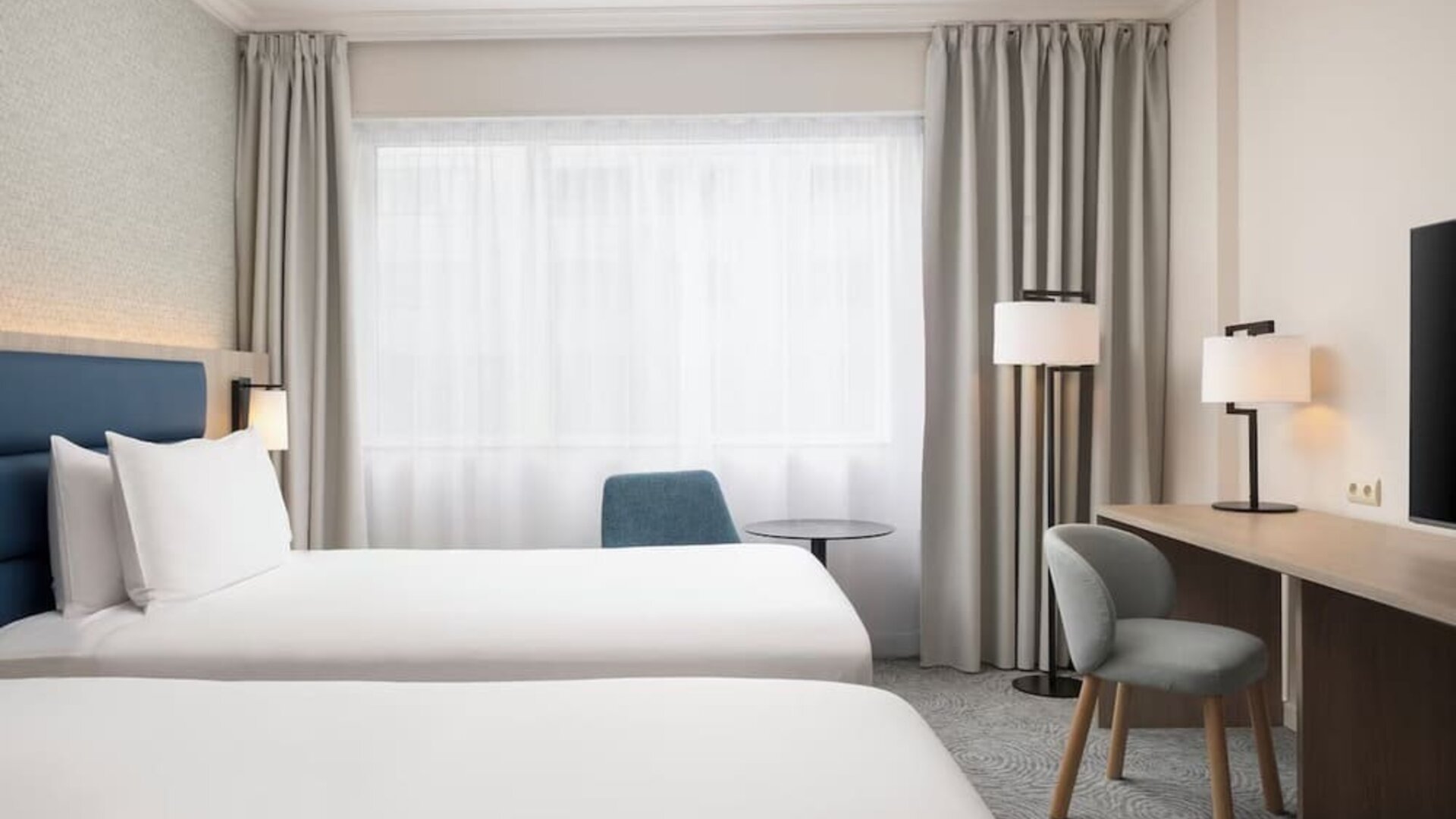 Bright hotel guest room with two beds, large window, desk, chairs, bedside lighting and wall-mounted television.