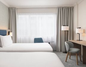 Bright hotel guest room with two beds, large window, desk, chairs, bedside lighting and wall-mounted television.