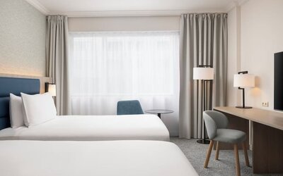 Bright hotel guest room with two beds, large window, desk, chairs, bedside lighting and wall-mounted television.