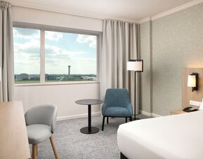 Spacious king guest room with large airport-view window, work desk, armchairs, side table and contemporary furnishings.