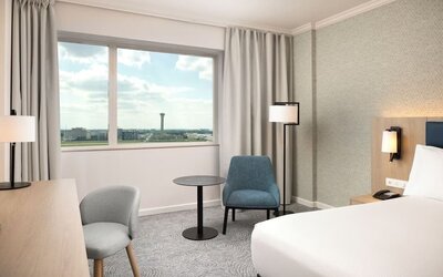 Spacious king guest room with large airport-view window, work desk, armchairs, side table and contemporary furnishings.