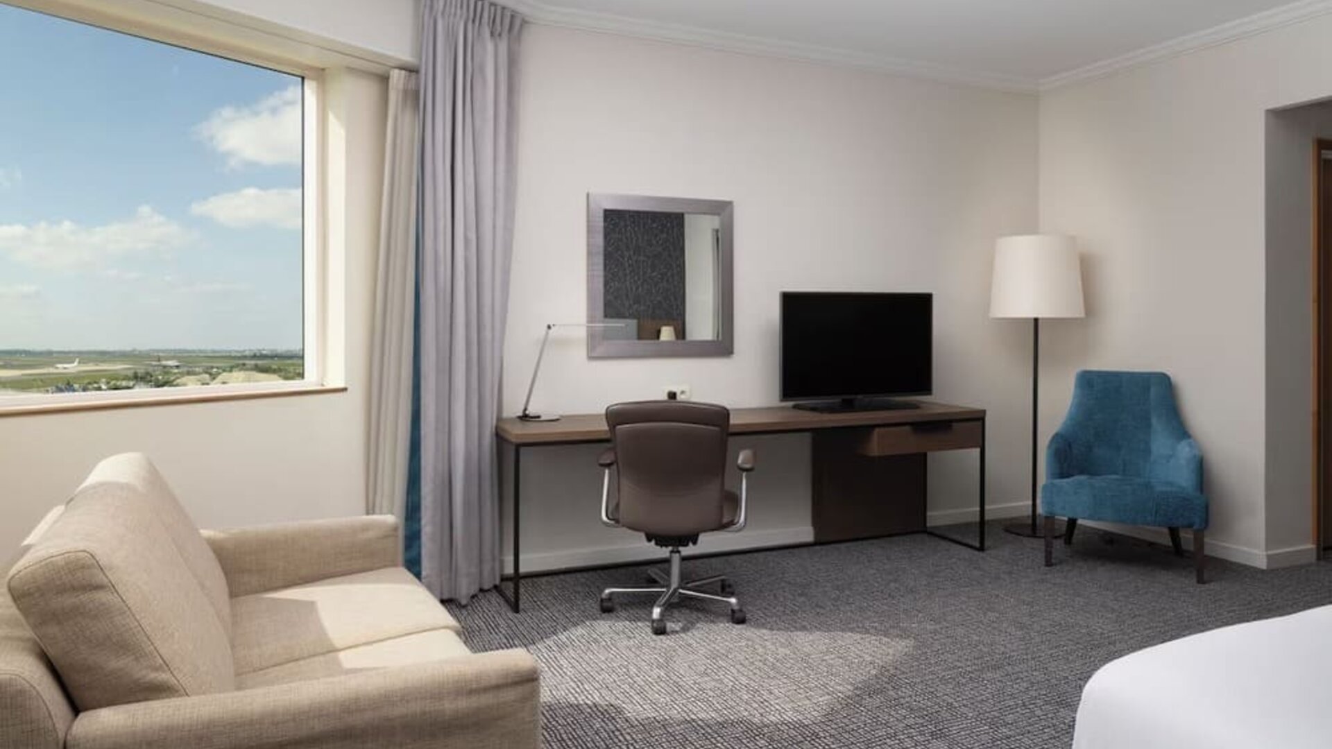Spacious guest room workspace with desk, office chair, television, sofa, armchair and large window overlooking the airport. 
