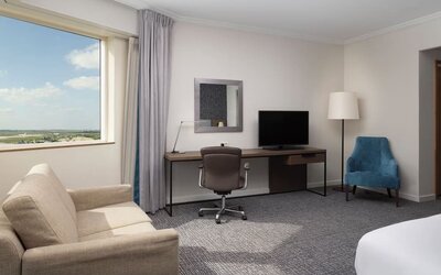 Spacious guest room workspace with desk, office chair, television, sofa, armchair and large window overlooking the airport. 