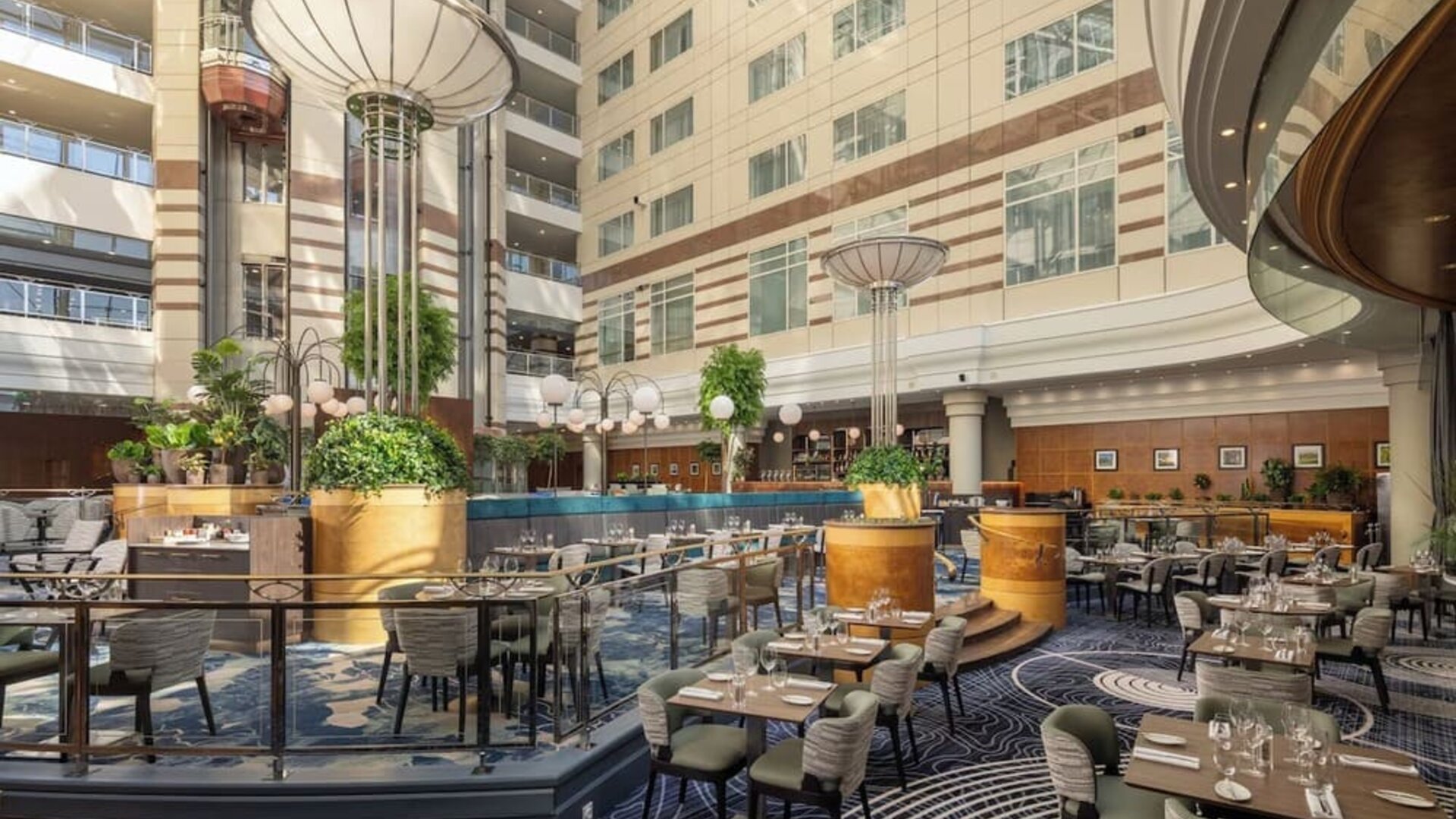 Large hotel atrium with restaurant tables, lounge seating, indoor trees, decorative columns and soaring glass ceiling