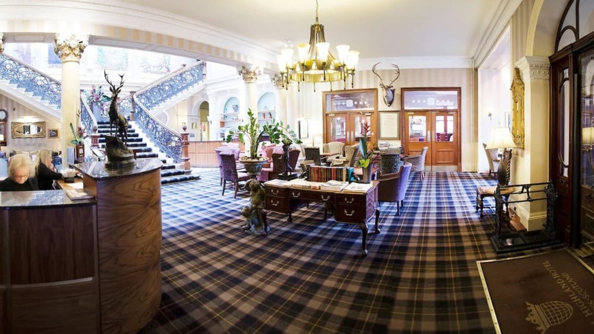 Grand hotel lobby featuring tartan carpeting, elegant seating, a sweeping staircase, and traditional Scottish décor at The Royal Highland Hotel.