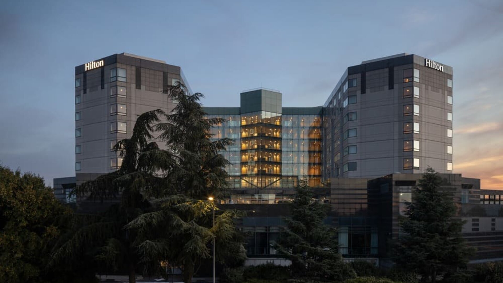 Hotel exterior at dusk with trees and garden setting. 