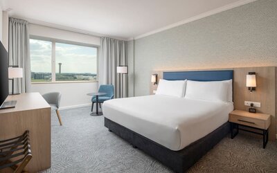 Modern king guest room with bed, large television, long work desk, armchairs, side table and floor-to-ceiling curtains.