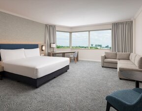 Spacious king guest room with large airport-view window, work desk, armchairs, side table and contemporary furnishings.