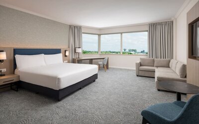 Spacious king guest room with large airport-view window, work desk, armchairs, side table and contemporary furnishings.