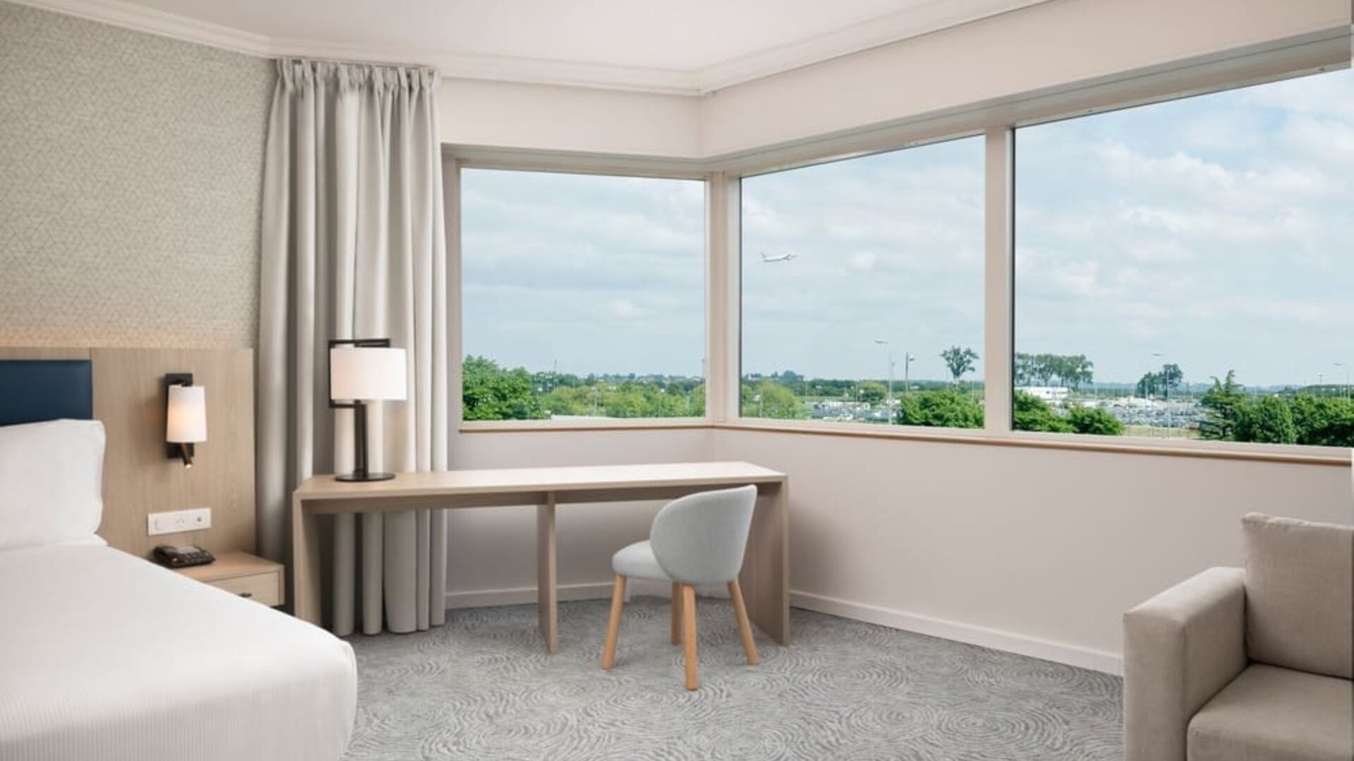 Spacious king guest room with large airport-view window, work desk, armchairs, side table and contemporary furnishings.
