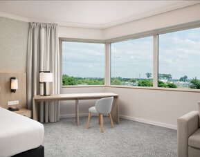 Spacious king guest room with large airport-view window, work desk, armchairs, side table and contemporary furnishings.
