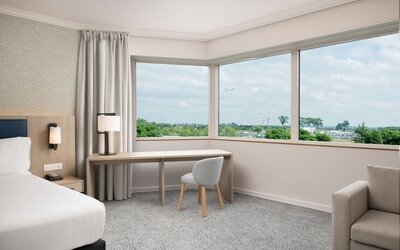 Spacious king guest room with large airport-view window, work desk, armchairs, side table and contemporary furnishings.