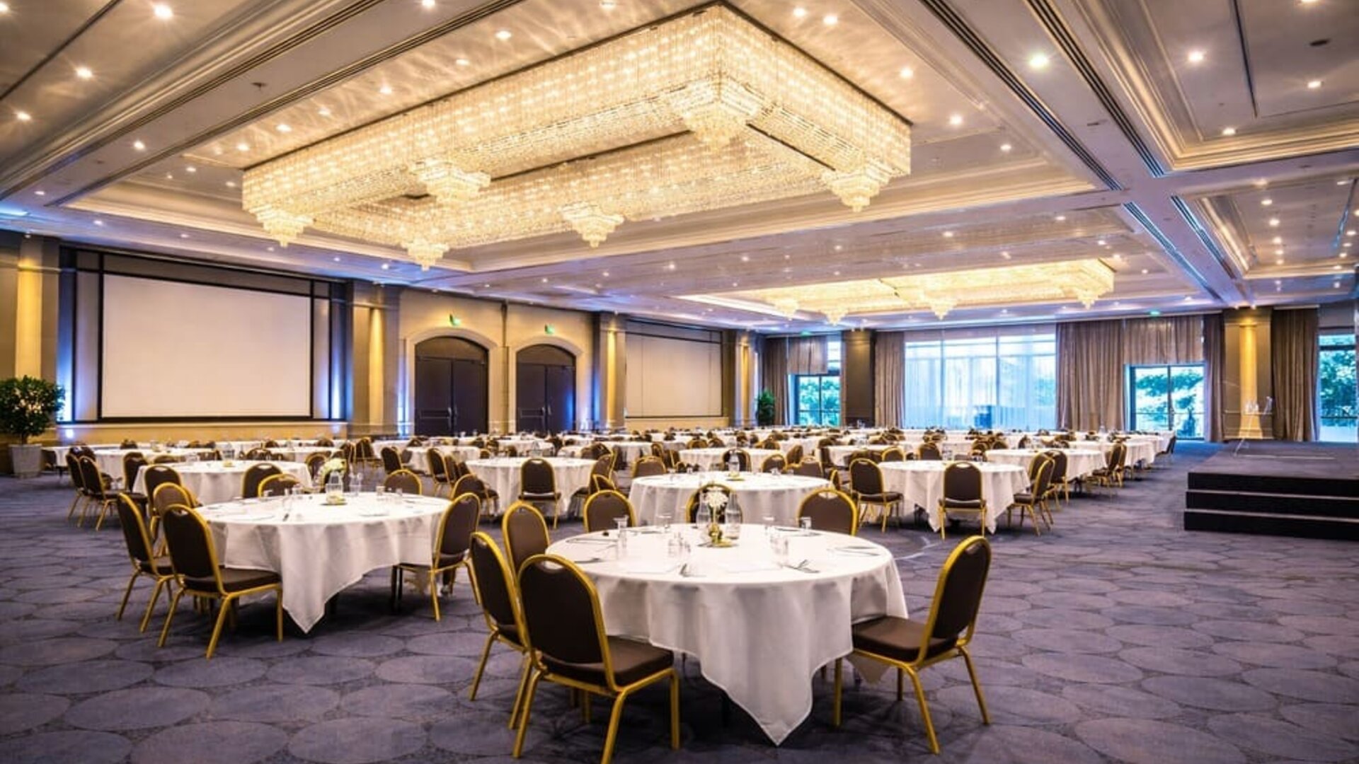 Hotel conference room with round conference tables, chairs, presentation screen, and natural light. 