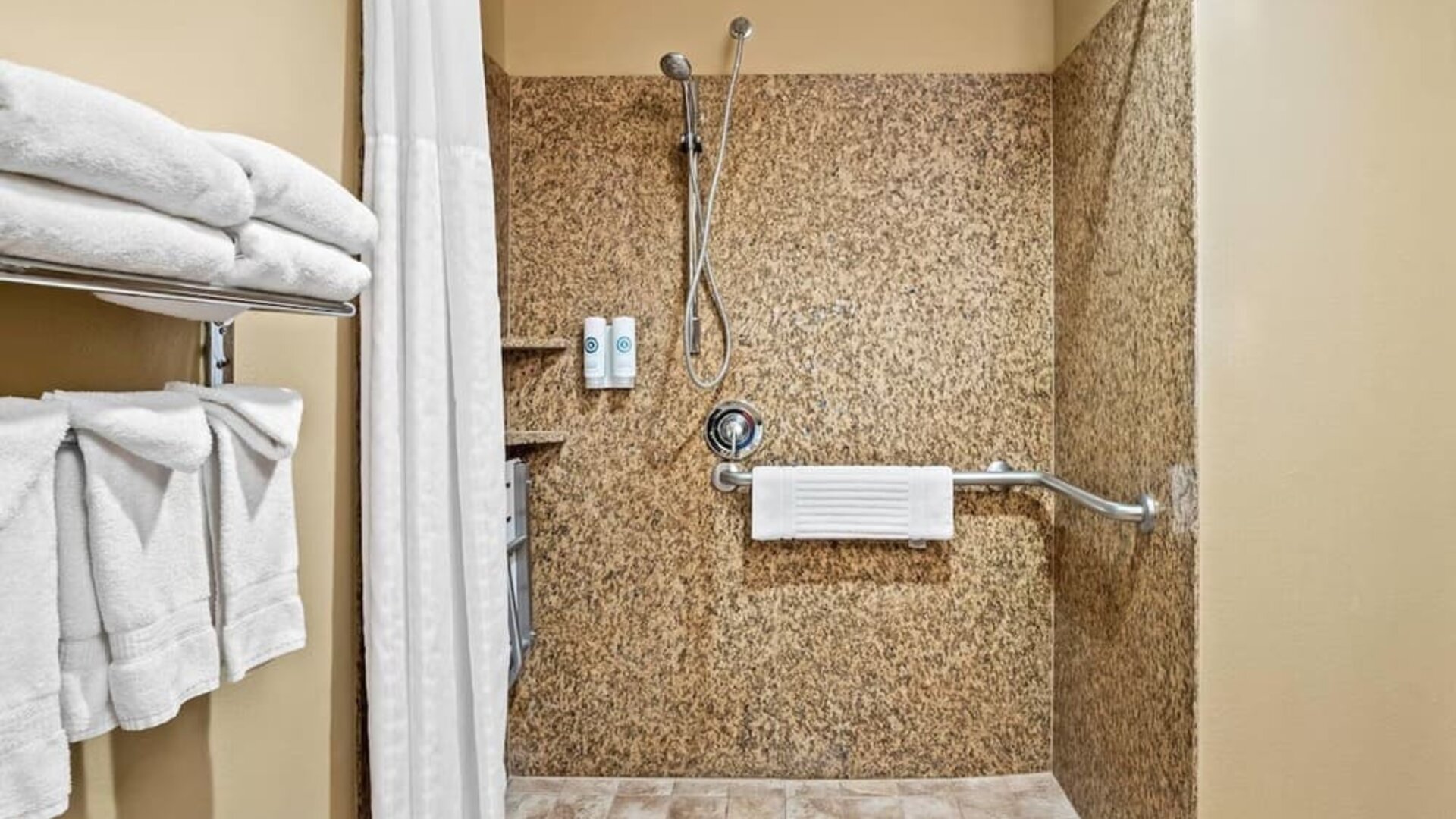 Accessible walk-in shower with granite-style walls, grab bars, mounted bath amenities, and neatly arranged towels at Comfort Suites Frisco.