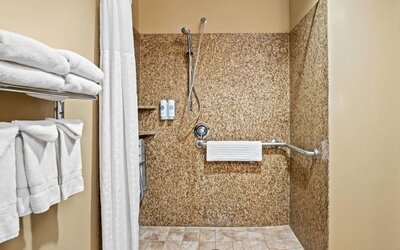 Accessible walk-in shower with granite-style walls, grab bars, mounted bath amenities, and neatly arranged towels at Comfort Suites Frisco.