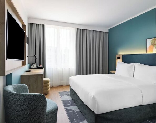 Bright contemporary hotel room with a king bed, white bedding, teal accent wall, large window with gray curtains, wall-mounted TV, desk, and upholstered armchair.