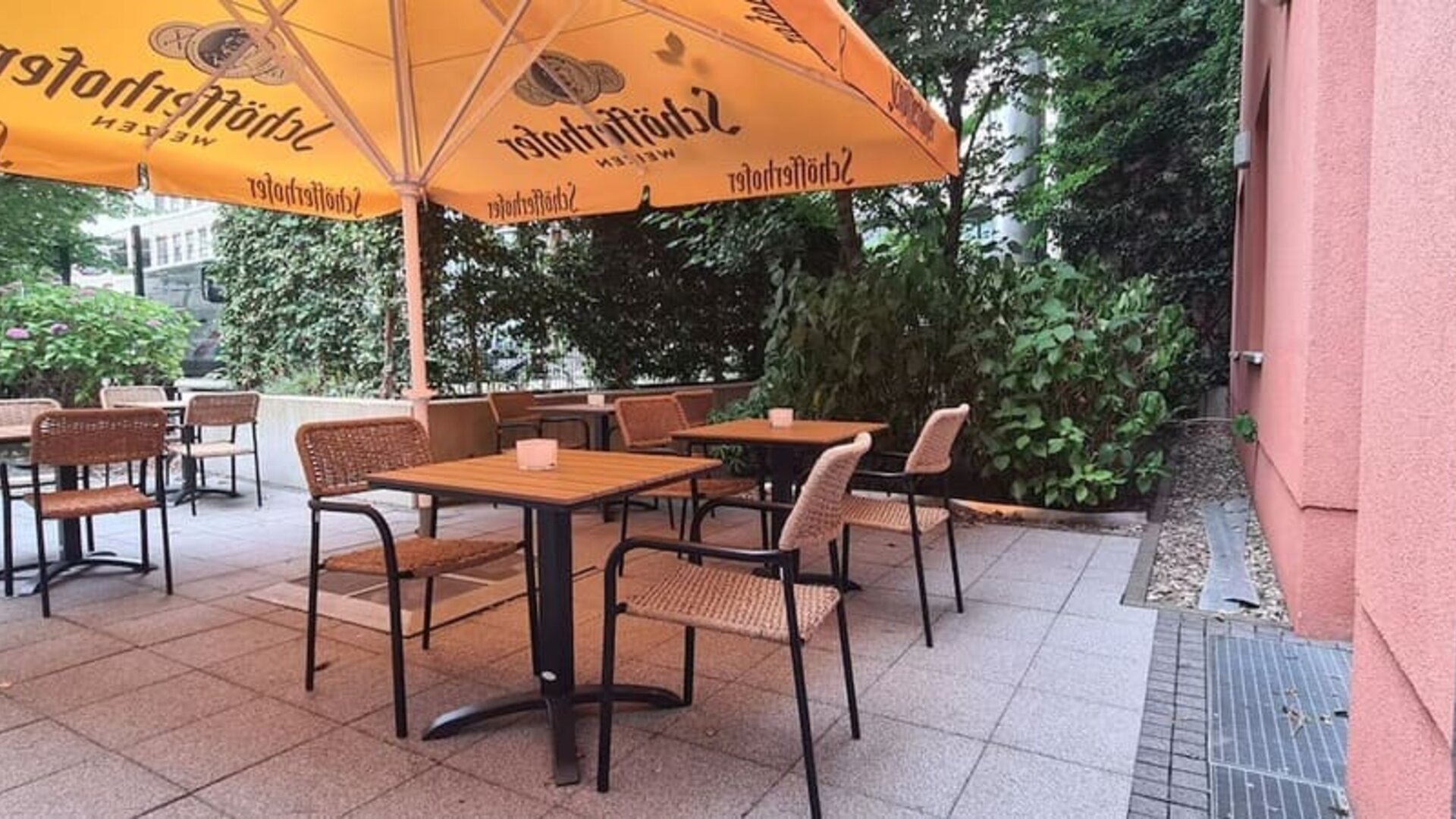 Shaded hotel terrace with wooden tables, woven chairs, large orange umbrellas, tiled flooring, and dense green trees and shrubs surrounding the seating area.