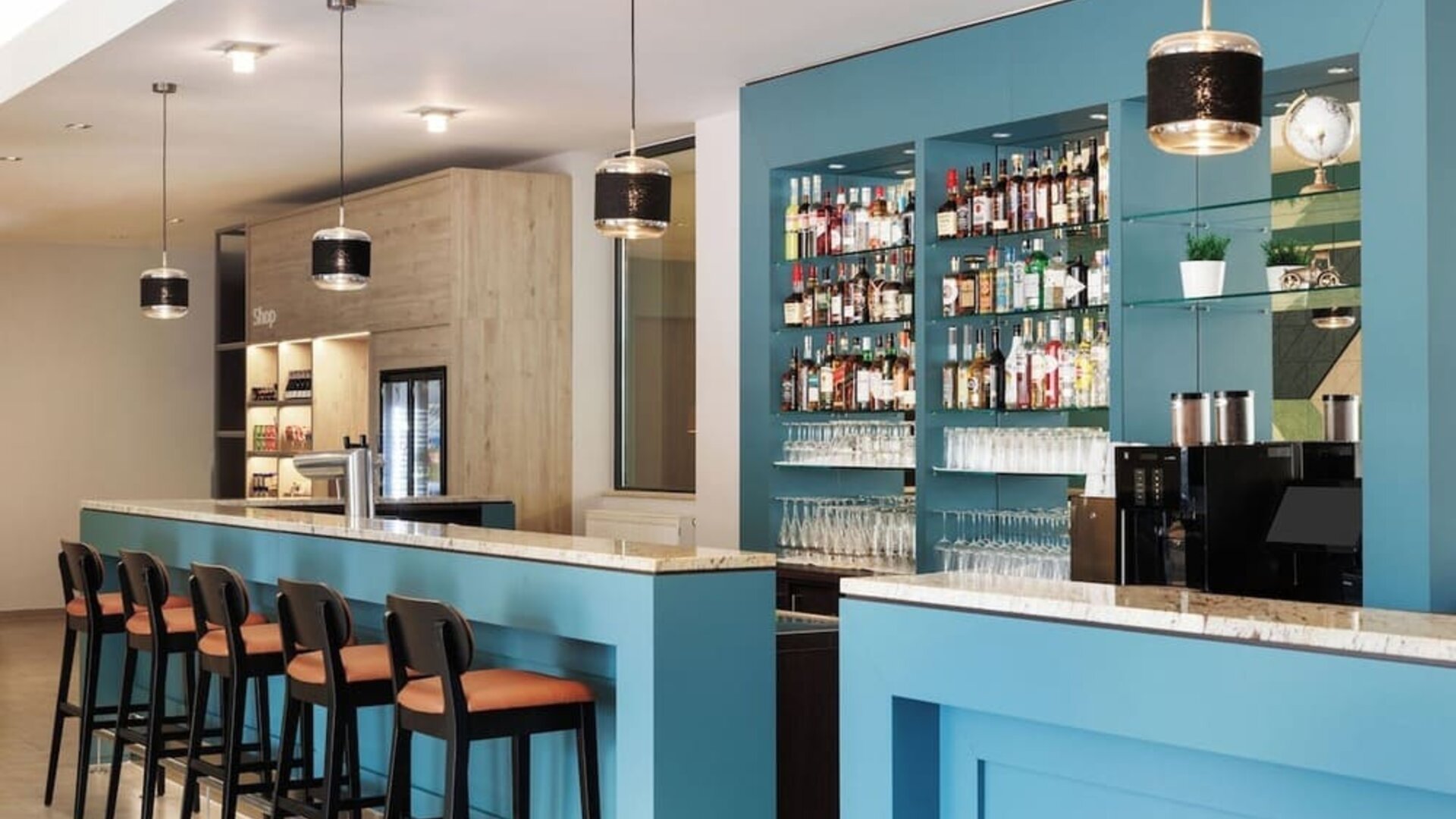 Contemporary hotel bar with turquoise counters, marble-look surfaces, high stools, pendant lighting, extensive bottle displays, glassware, and a coffee machine.