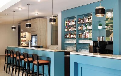Contemporary hotel bar with turquoise counters, marble-look surfaces, high stools, pendant lighting, extensive bottle displays, glassware, and a coffee machine.