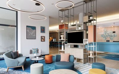 Contemporary hotel lobby with colorful lounge chairs and ottomans, round tables, blue patterned carpeting, circular ceiling lights, open shelving, and reception desk in the background.