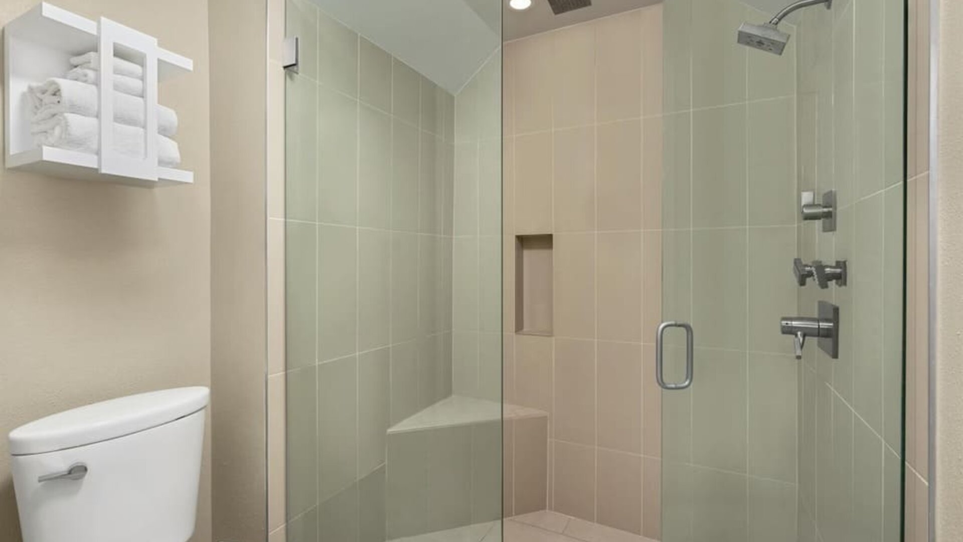 Modern hotel bathroom with a glass-enclosed walk-in shower, tiled walls, rainfall-style showerhead, built-in corner seat and wall niche, toilet, and open shelving with folded towels.