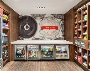 Hotel convenience shop with wooden shelving stocked with snacks, refrigerated cases containing bottled and canned drinks, and a central counter beneath a large “Treats” wall graphic.