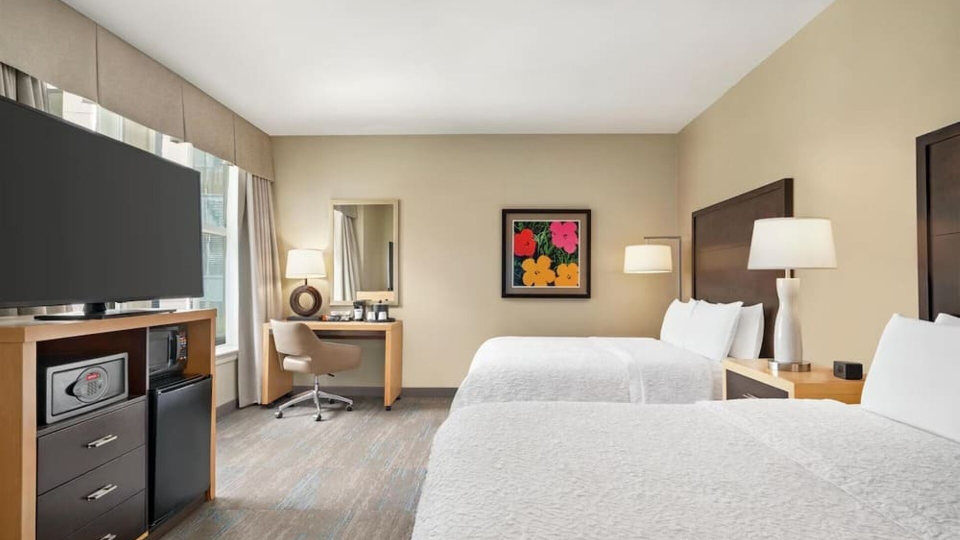 Spacious hotel guest room with two beds dressed in white linens, dark wood headboards, bedside lamps, desk and chair, flat-screen TV, microwave, mini refrigerator, and colorful framed artwork.