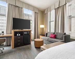 Bright corner hotel room with large windows, gray sofa with pink accent pillow, desk and chair, ottoman, flat-screen TV, microwave, mini refrigerator, and part of a bed visible.