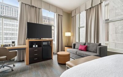 Bright corner hotel room with large windows, gray sofa with pink accent pillow, desk and chair, ottoman, flat-screen TV, microwave, mini refrigerator, and part of a bed visible.