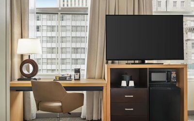 Guest room workspace with a desk and swivel chair beside a large window, flat-screen TV, microwave, mini refrigerator, storage drawers, coffee supplies, and decorative table lamp.