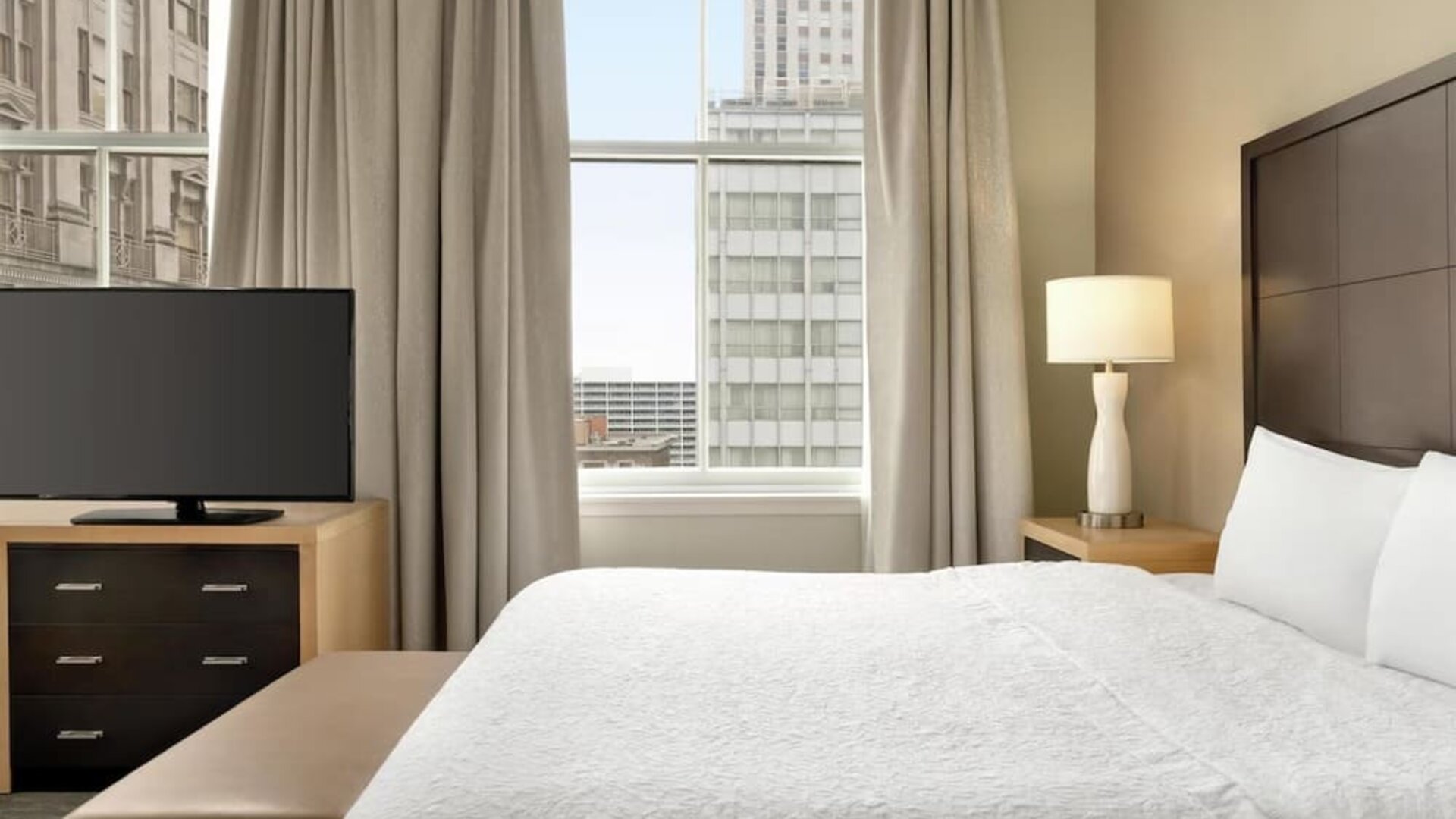 Hotel guest room with a king bed, white bedding, dark wood headboard, bedside lamp, flat-screen TV on a dresser, upholstered bench, and tall window overlooking surrounding city buildings.