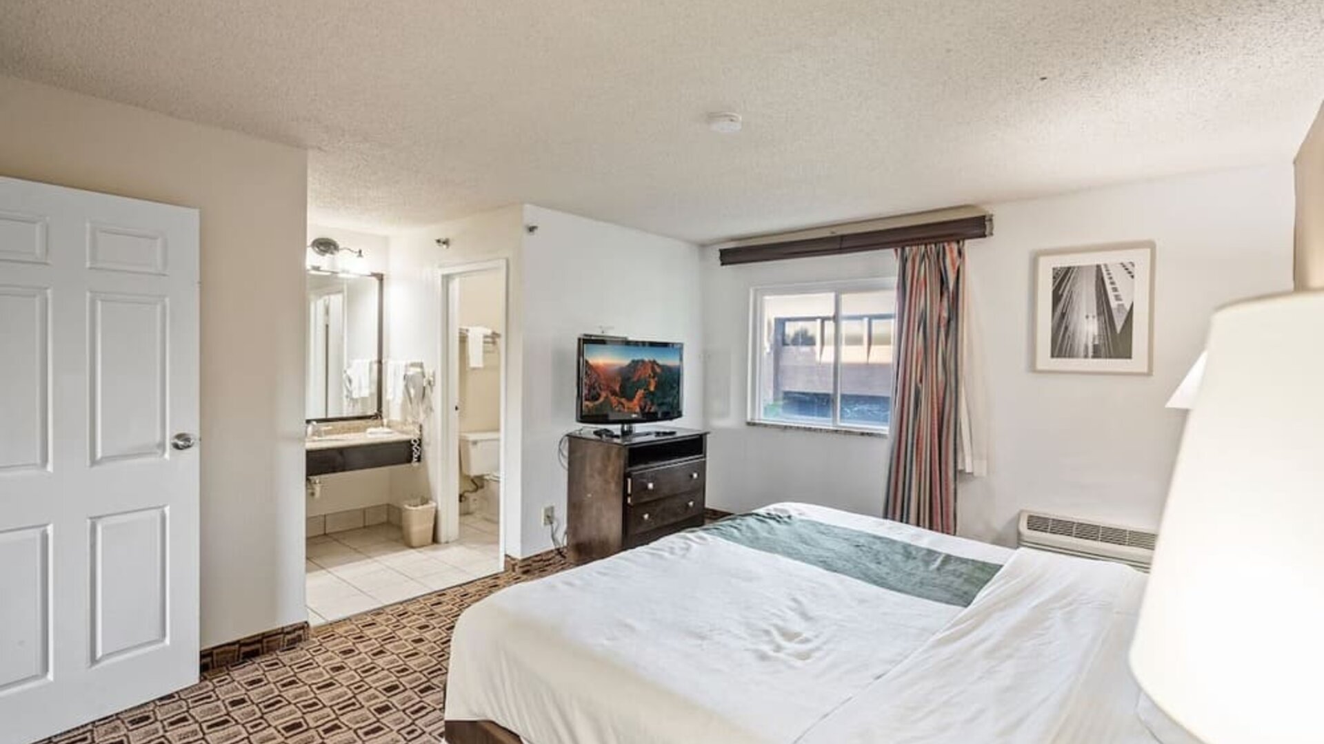 Spacious king guest room featuring a king bed, flat-screen television, large window, and convenient access to the bathroom at Coratel Plus Suites Wichita West Airport.