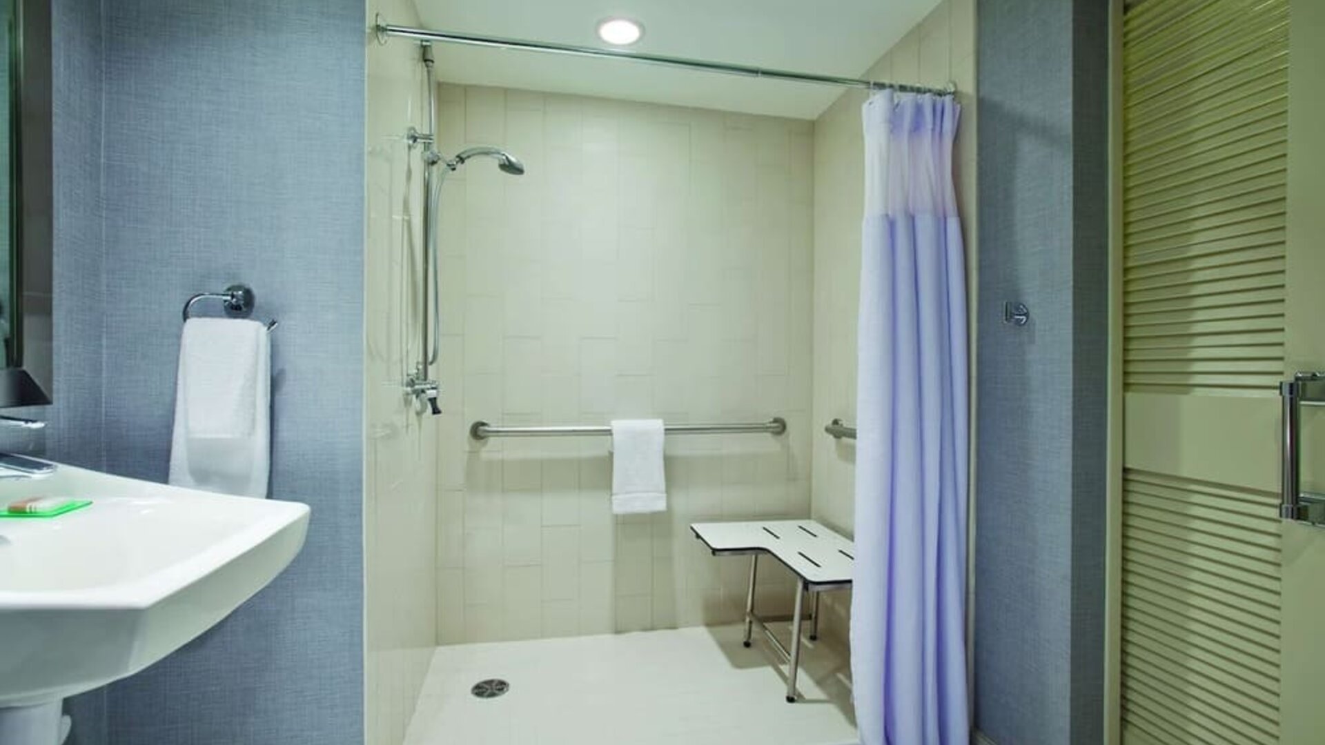 Accessible guest bathroom featuring a roll-in shower, grab bars, fold-down shower seat, and pedestal sink at Hyatt Place San Jose Downtown.