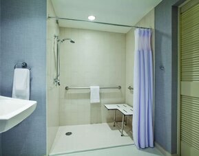 Accessible guest bathroom featuring a roll-in shower, grab bars, fold-down shower seat, and pedestal sink at Hyatt Place San Jose Downtown.