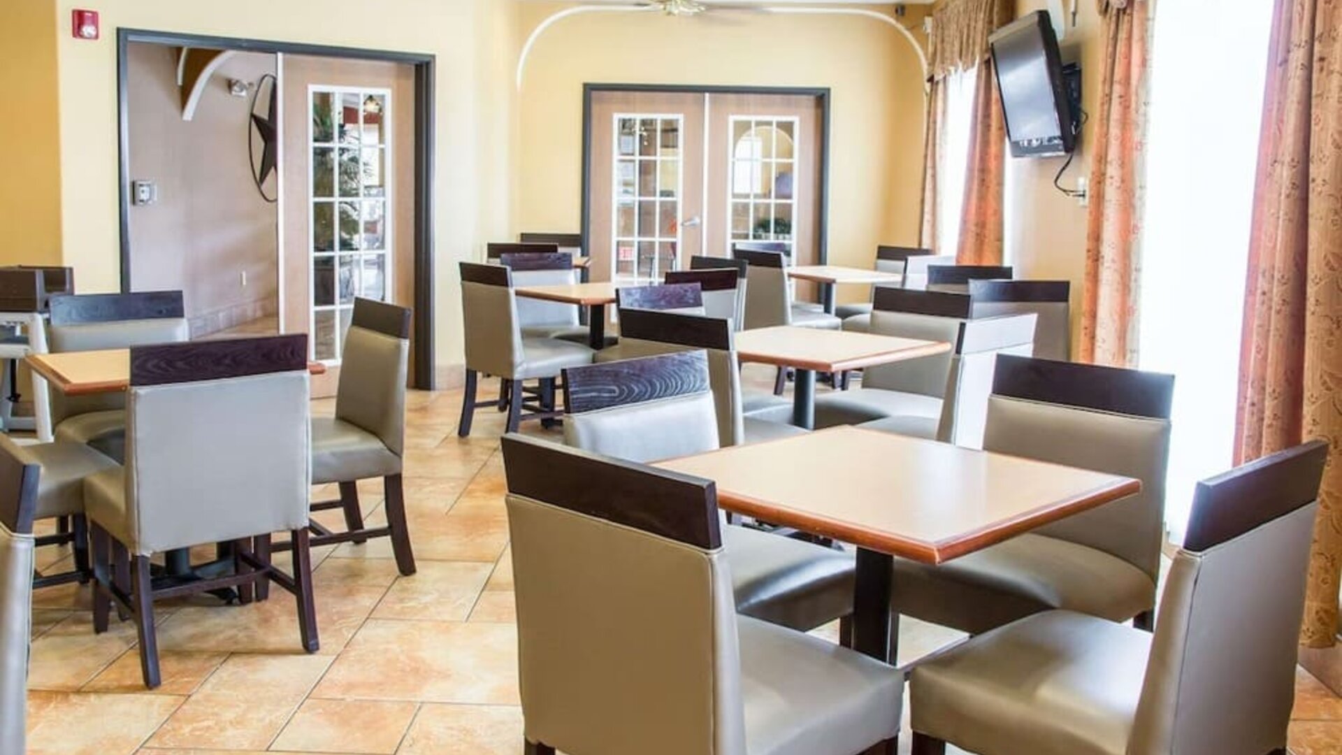 Bright hotel dining area with multiple tables, chairs, television, and large windows at Comfort Suites San Antonio SeaWorld - Lackland AFB.