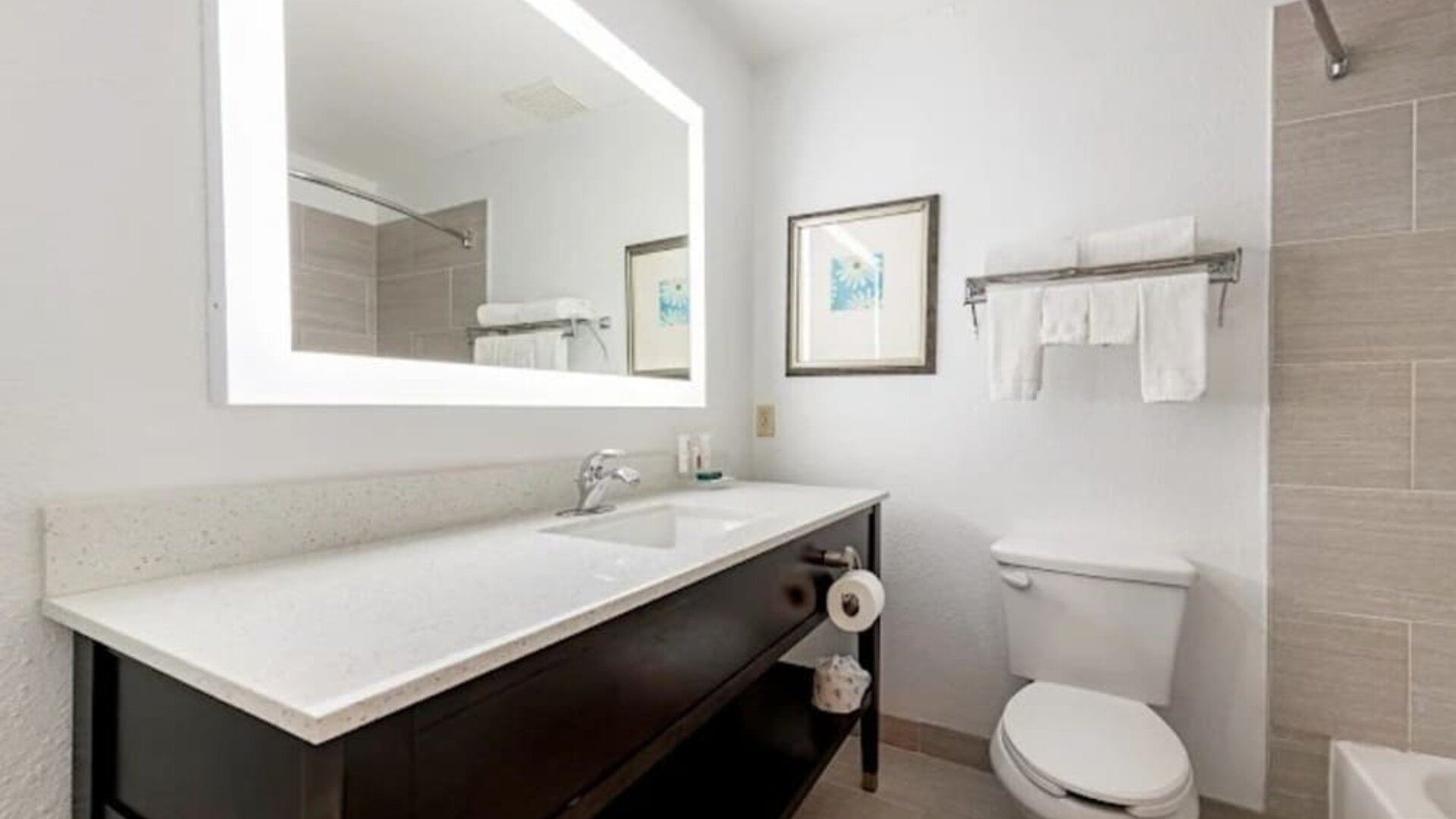 Guest bathroom with illuminated mirror, vanity, toilet, towels, and tiled bath area at SureStay by Best Western Lewisville Dallas.