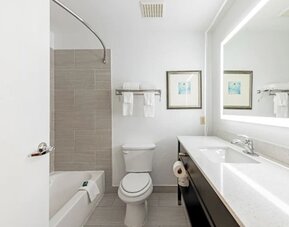 Guest bathroom with bathtub, tiled shower wall, toilet, long vanity, and illuminated mirror at SureStay by Best Western Lewisville Dallas.