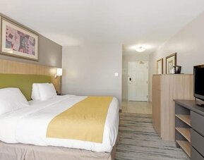 King guest room with white bedding, yellow bed runner, green upholstered headboard, television, dresser, and storage cabinet at SureStay by Best Western Lewisville Dallas.