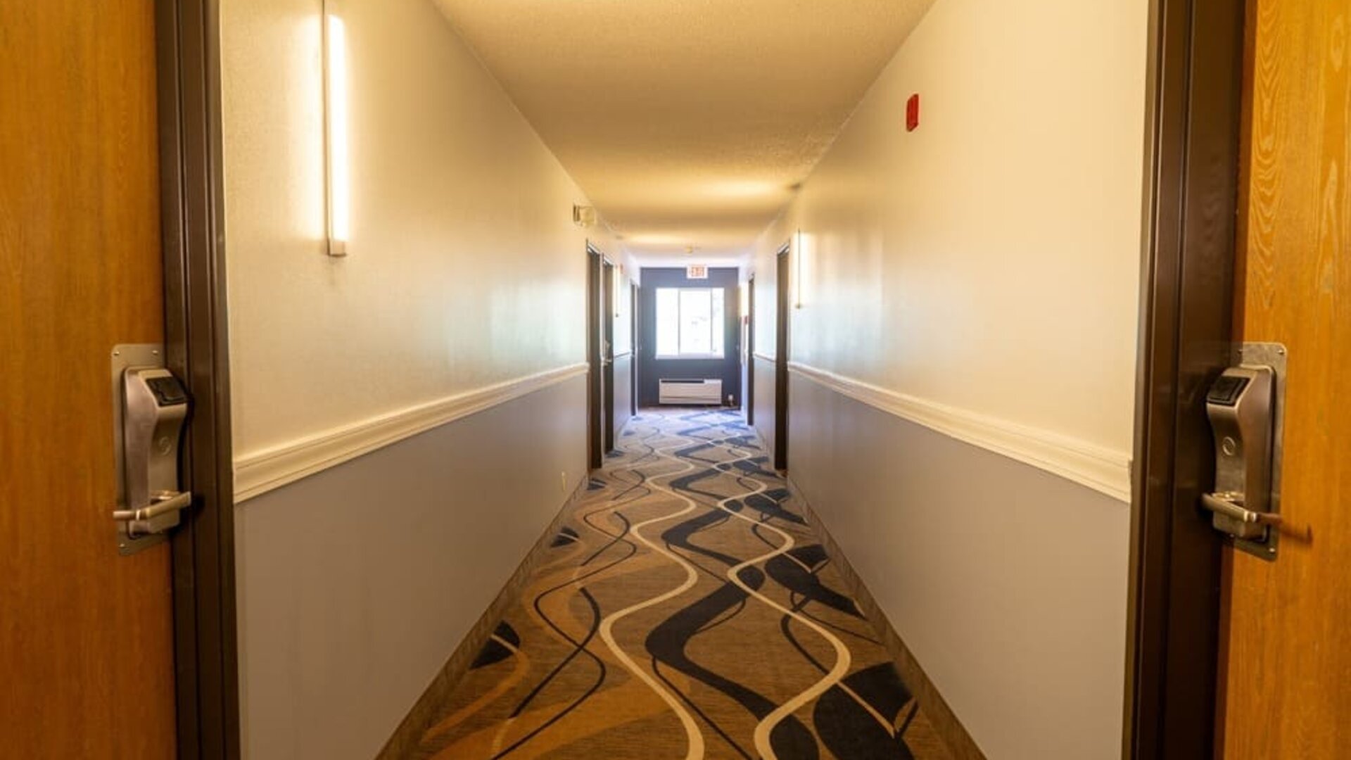 Hotel corridor with patterned carpeting, guest room doors, wall lighting, and window at the end of the hallway at Coratel Inn & Suites By Jasper Hastings.