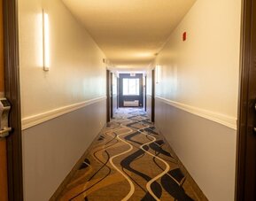 Hotel corridor with patterned carpeting, guest room doors, wall lighting, and window at the end of the hallway at Coratel Inn & Suites By Jasper Hastings.
