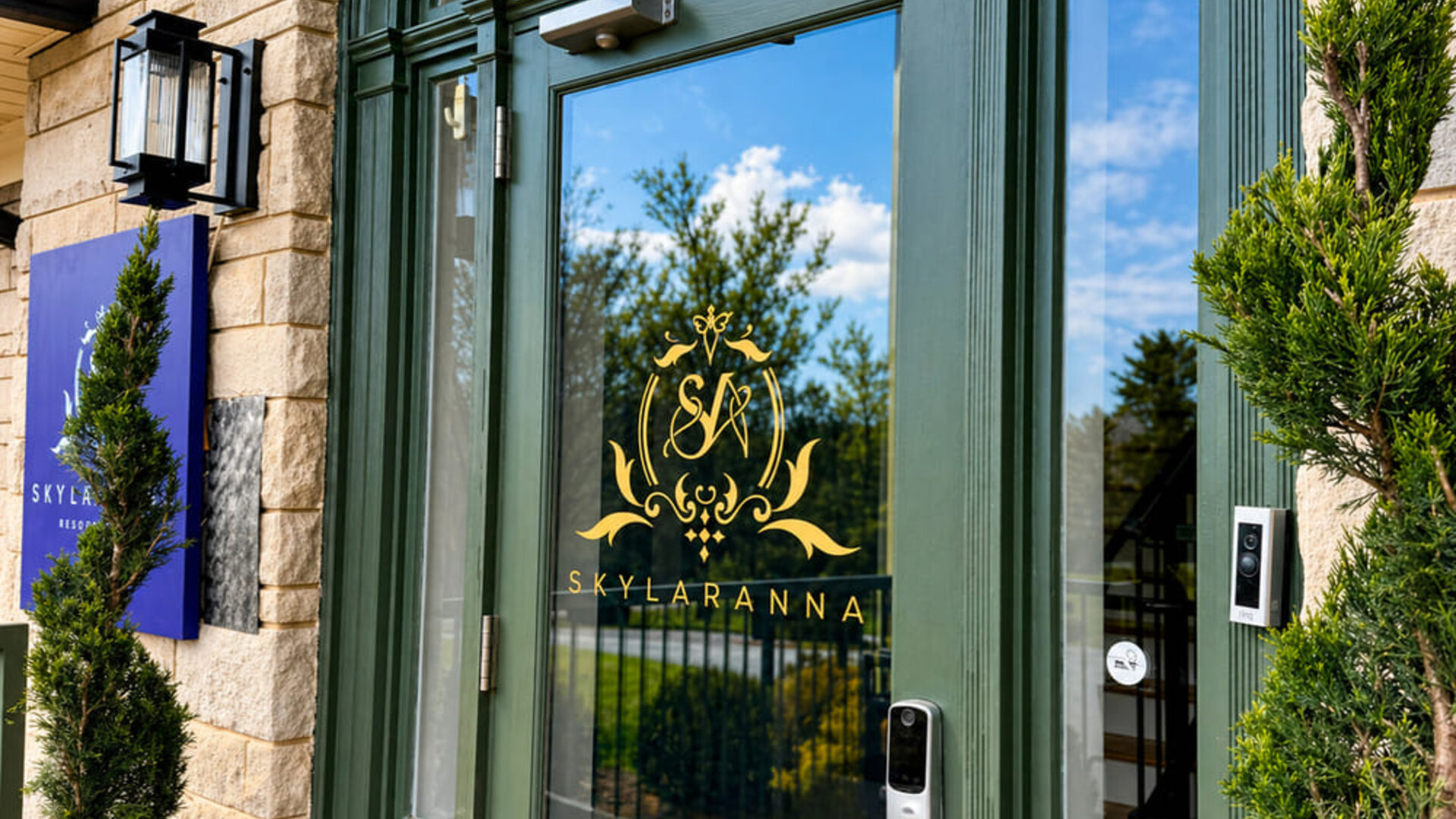 Main entrance to SKYLARANNA Resort & Spa, featuring glass doors with the resort logo, stone exterior, and decorative greenery.